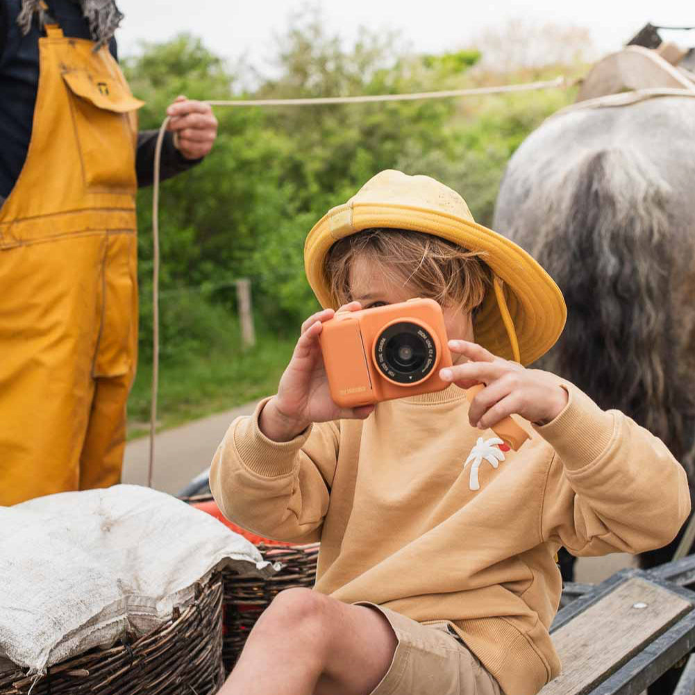 Child in yellow hat and overalls holding a camera, with an adult in yellow overalls standing nearby.