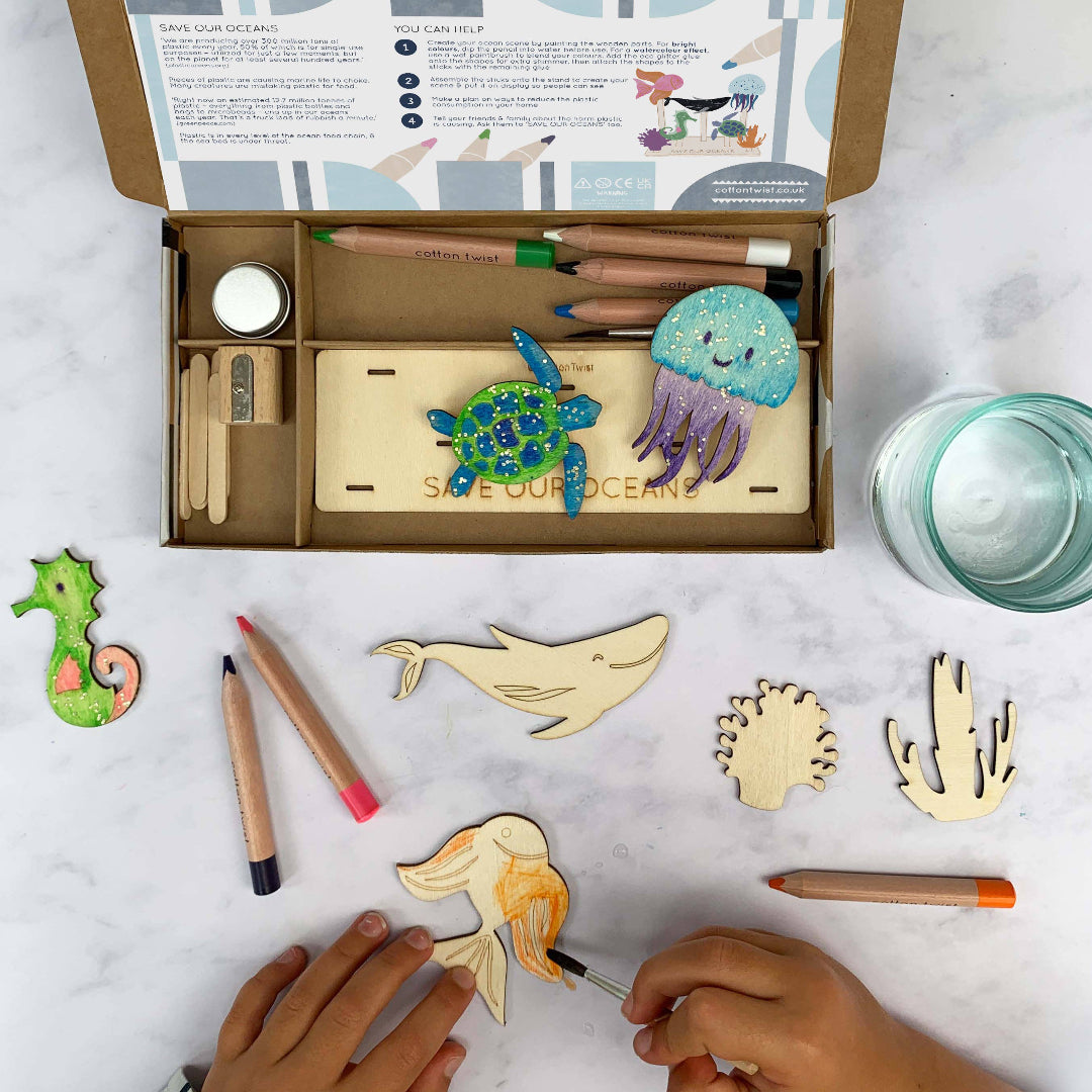 Children's craft kit with wooden ocean-themed shapes and coloring supplies on a marble surface.