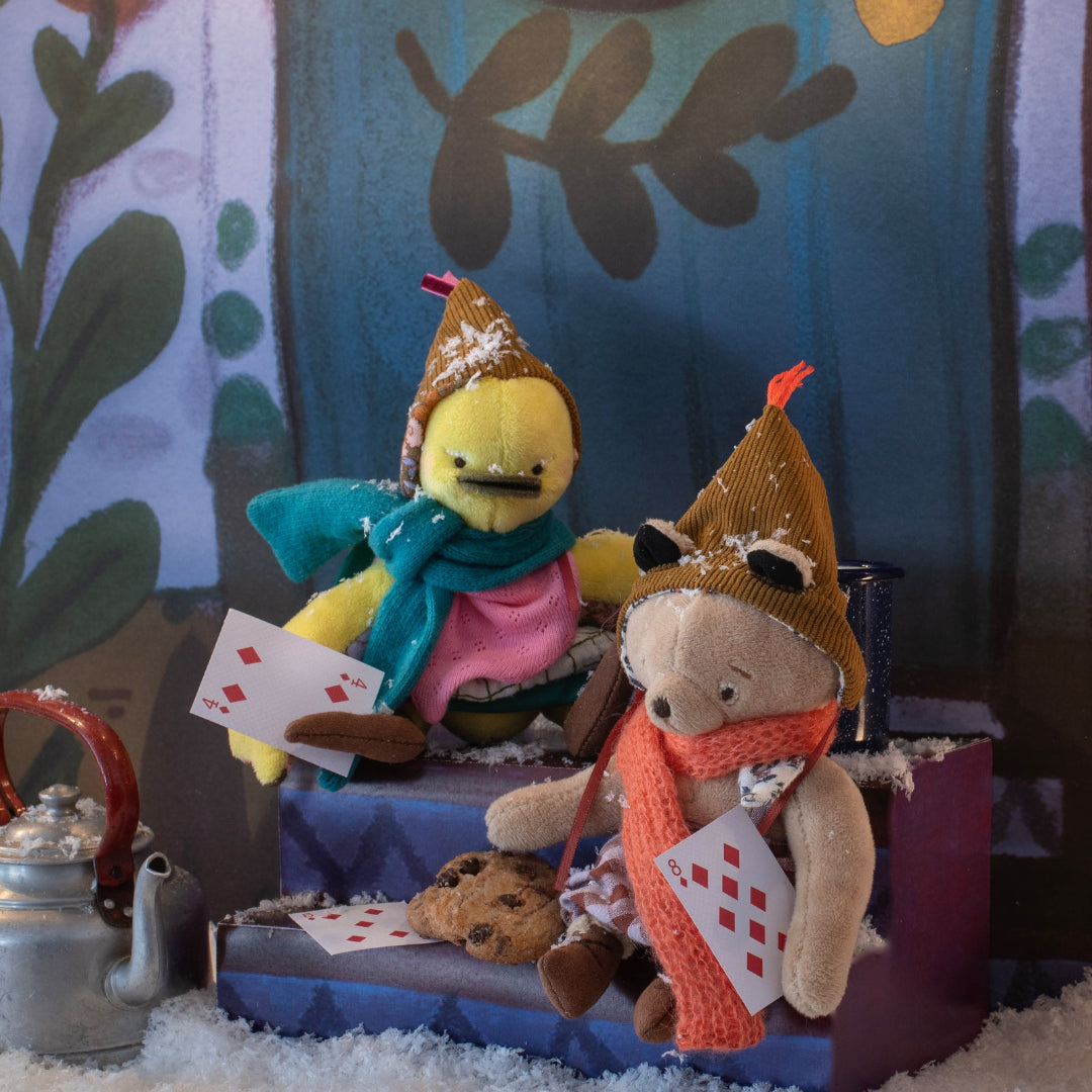 Two plush toys, a bird and a bear, sitting on a table with cards and cookies in a cozy setting.