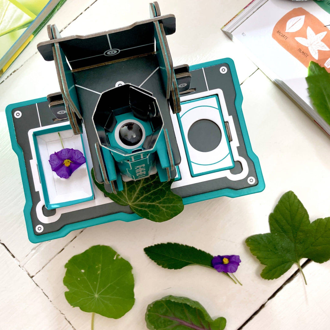 Microscope, surrounded by leaves and a book on a white surface.