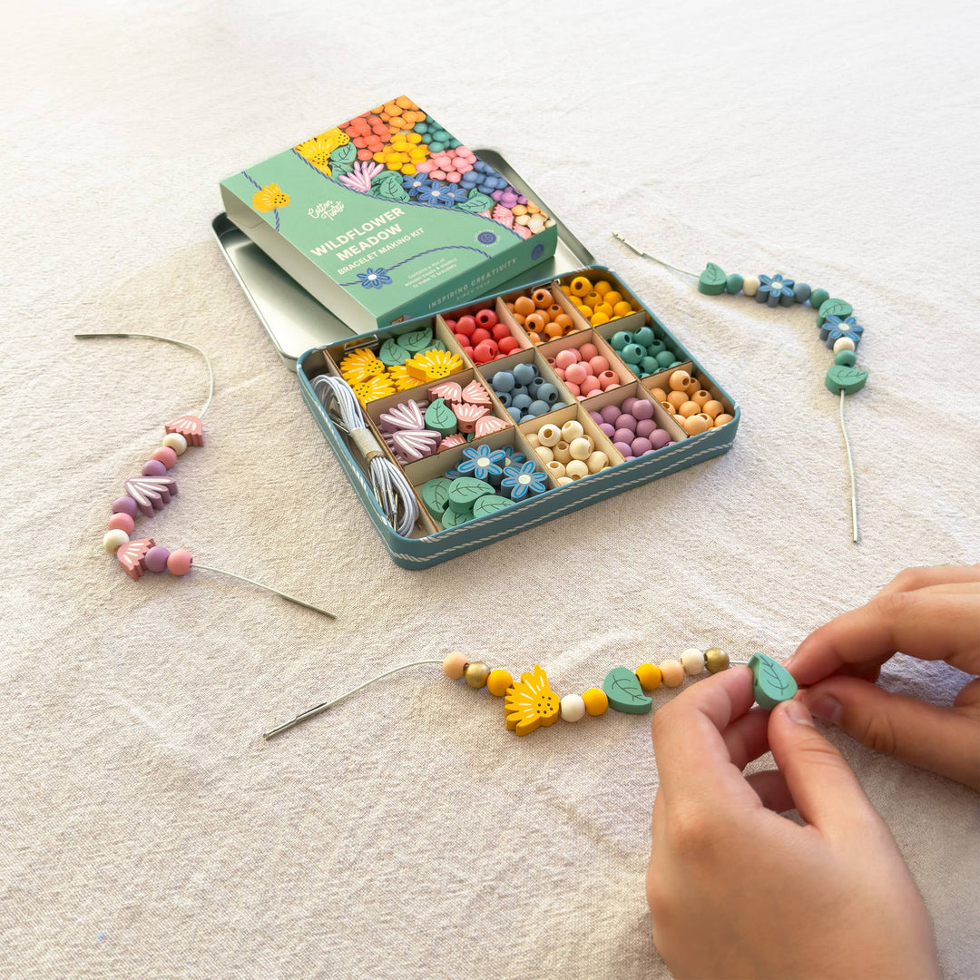 Child threading wooden flower and leaf beads onto a bracelet next to the open bead tin on a linen background