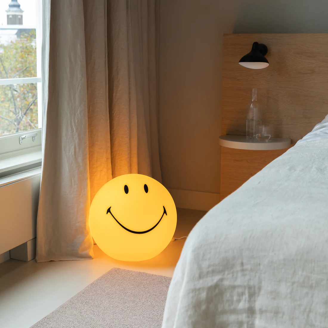 Smiley face night light on a bed in a bedroom with a window and lamp in the background.