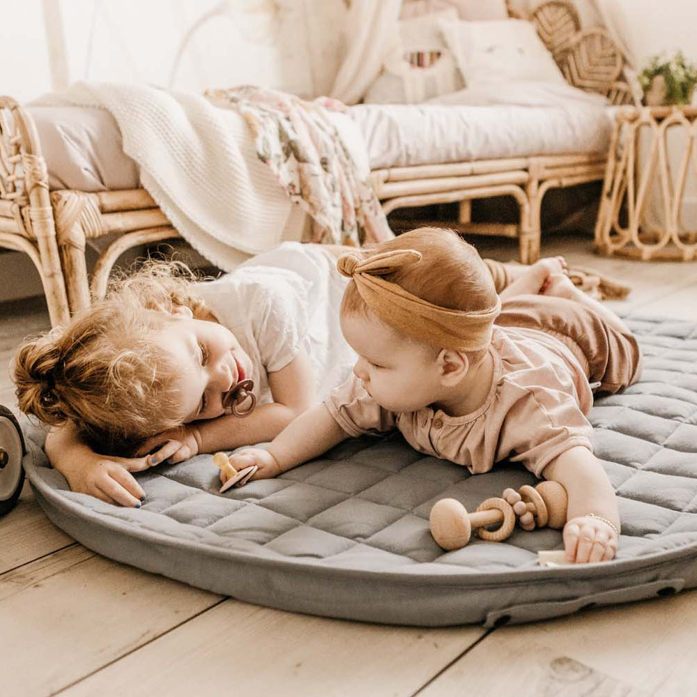 Two young children playing on a round gray mat in a cozy room with wicker furniture and soft lighting.
