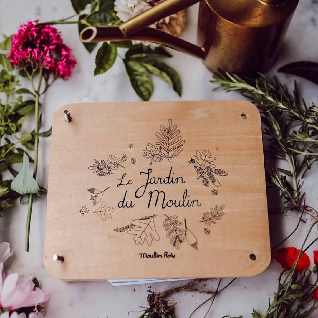 A square light wood flower press with 'Le Jardin du Moulin' and botanical illustrations printed on its front, styled on a marble surface with flowers, greenery, and a brass watering can.