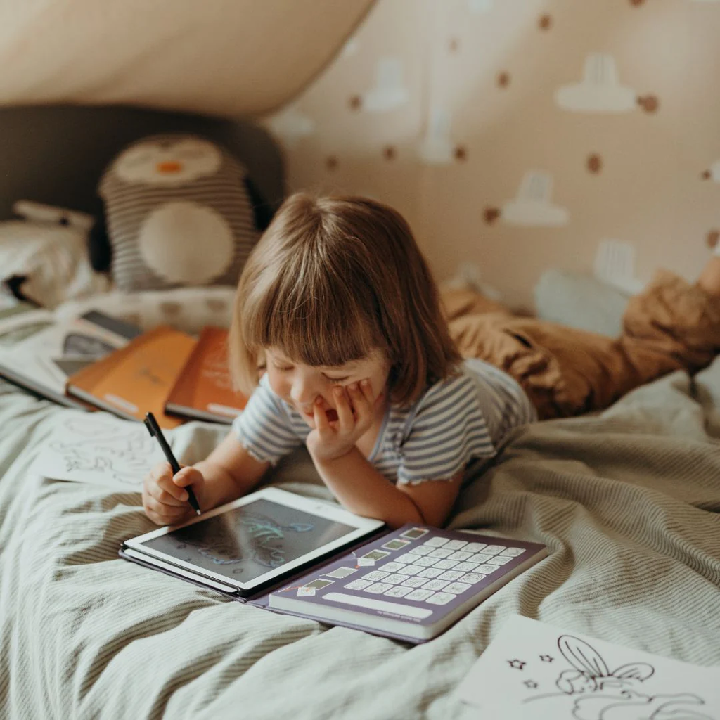 Child drawing in a notebook on a bed with a teddy bear and books around
