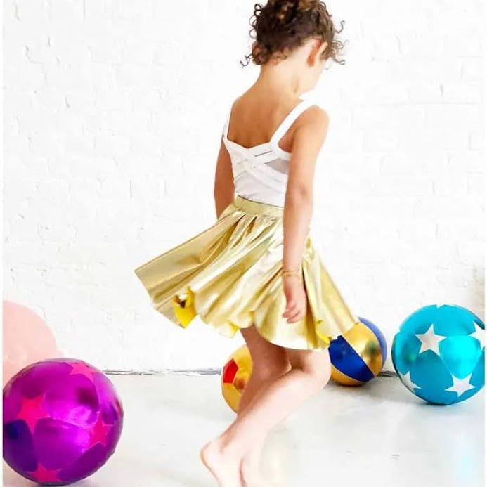Child in a gold skirt standing next to colorful balls on a white floor.