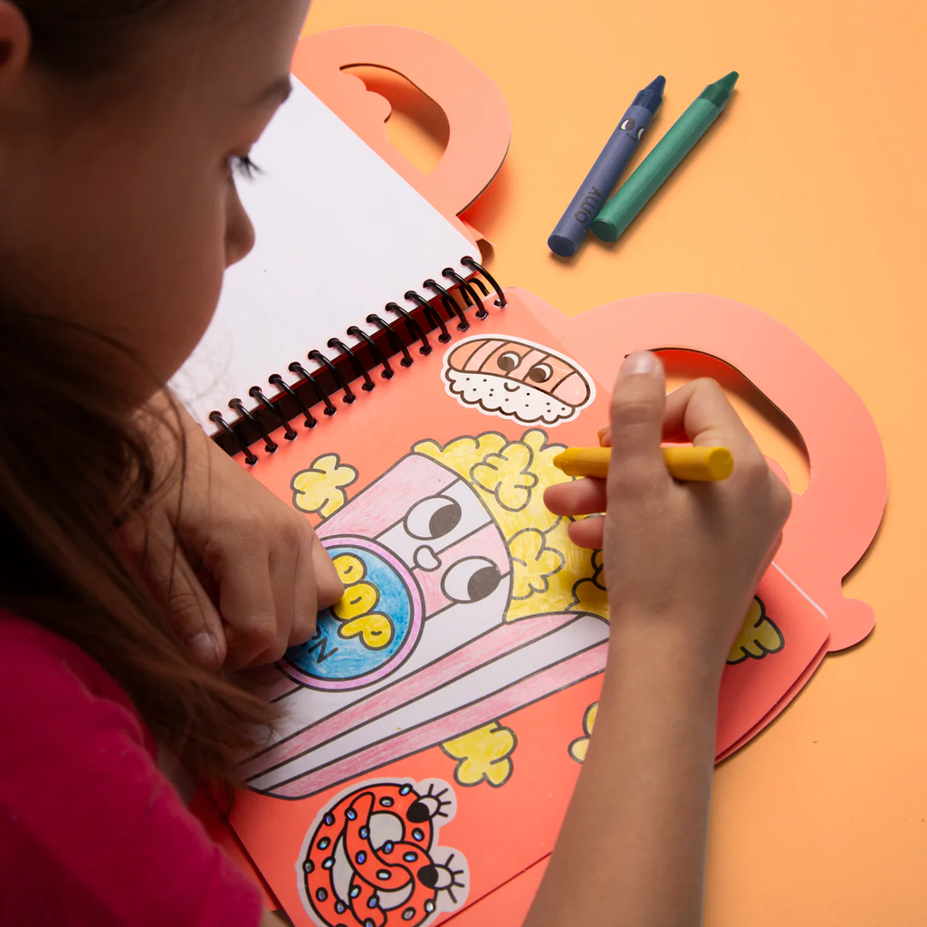Child coloring a cartoon character in a spiral-bound notebook with crayons on an orange background
