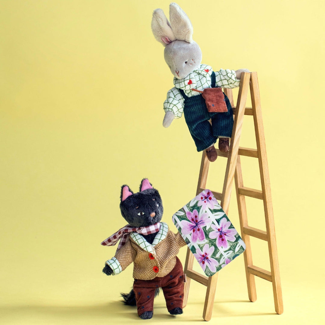 Two cloth toy animals posed with a small wooden ladder against a pale yellow studio background. The lower black cat-like doll wears a beige vest, brown pants, and a plaid neck scarf while holding a floral blanket.