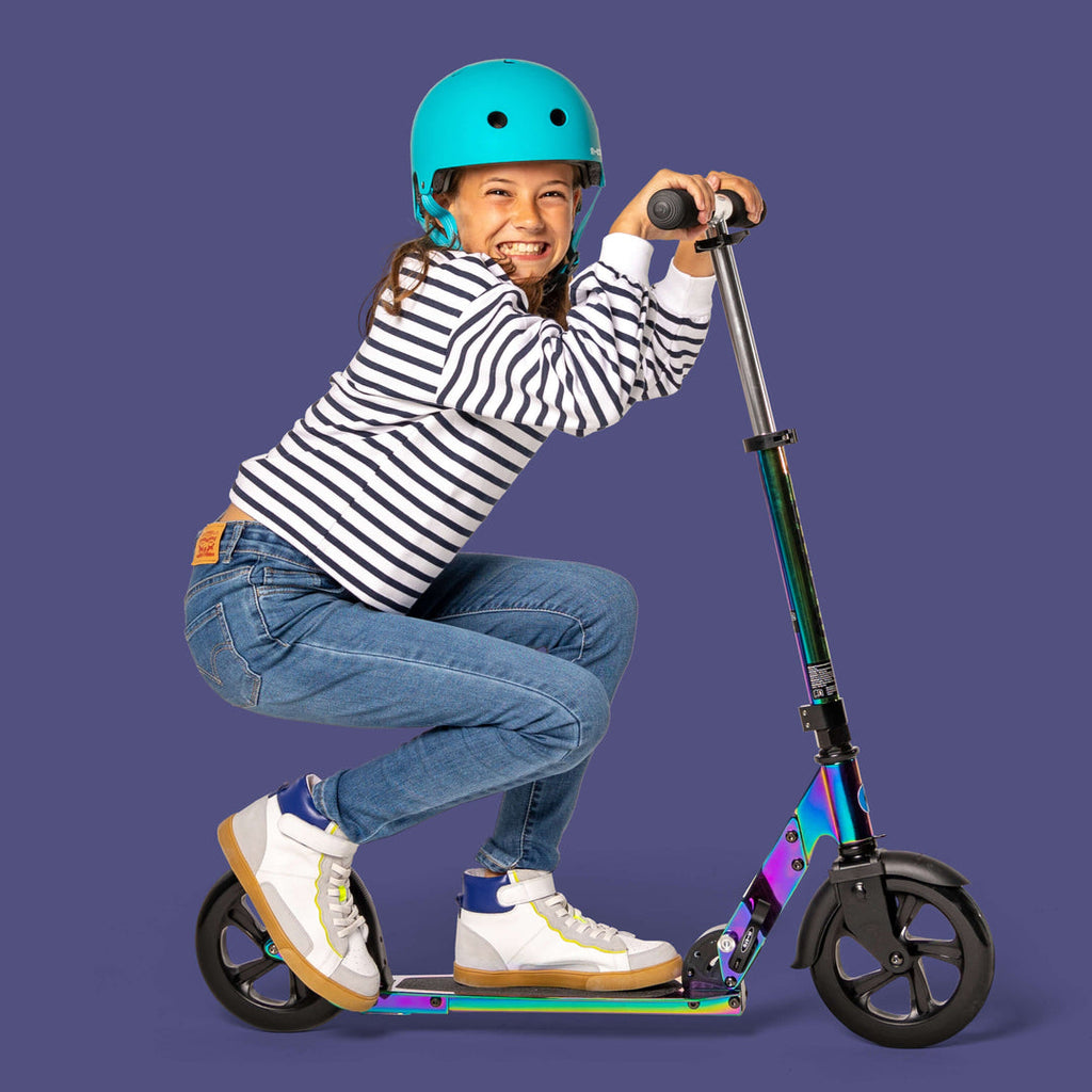 Laughing girl in a striped top wearing the aqua Micro helmet, crouched on a big-wheel scooter against a purple background