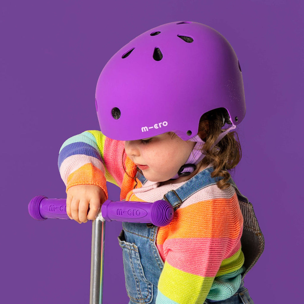 Toddler in a rainbow jumper wearing the purple Micro helmet and holding scooter handlebars against a purple background
