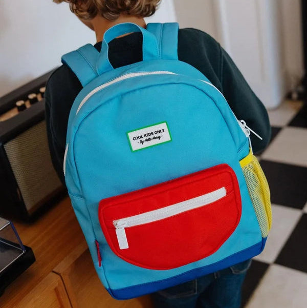A child wearing a bright turquoise backpack with a large red front pocket and a white zippered pocket, standing indoors near a wooden table and black-and-white checkered floor.